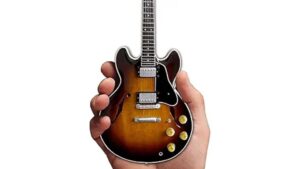 A person holds a miniature guitar-shaped phone case with detailed design and vibrant coloring, showcasing a unique tech accessory for music lovers and guitar enthusiasts.